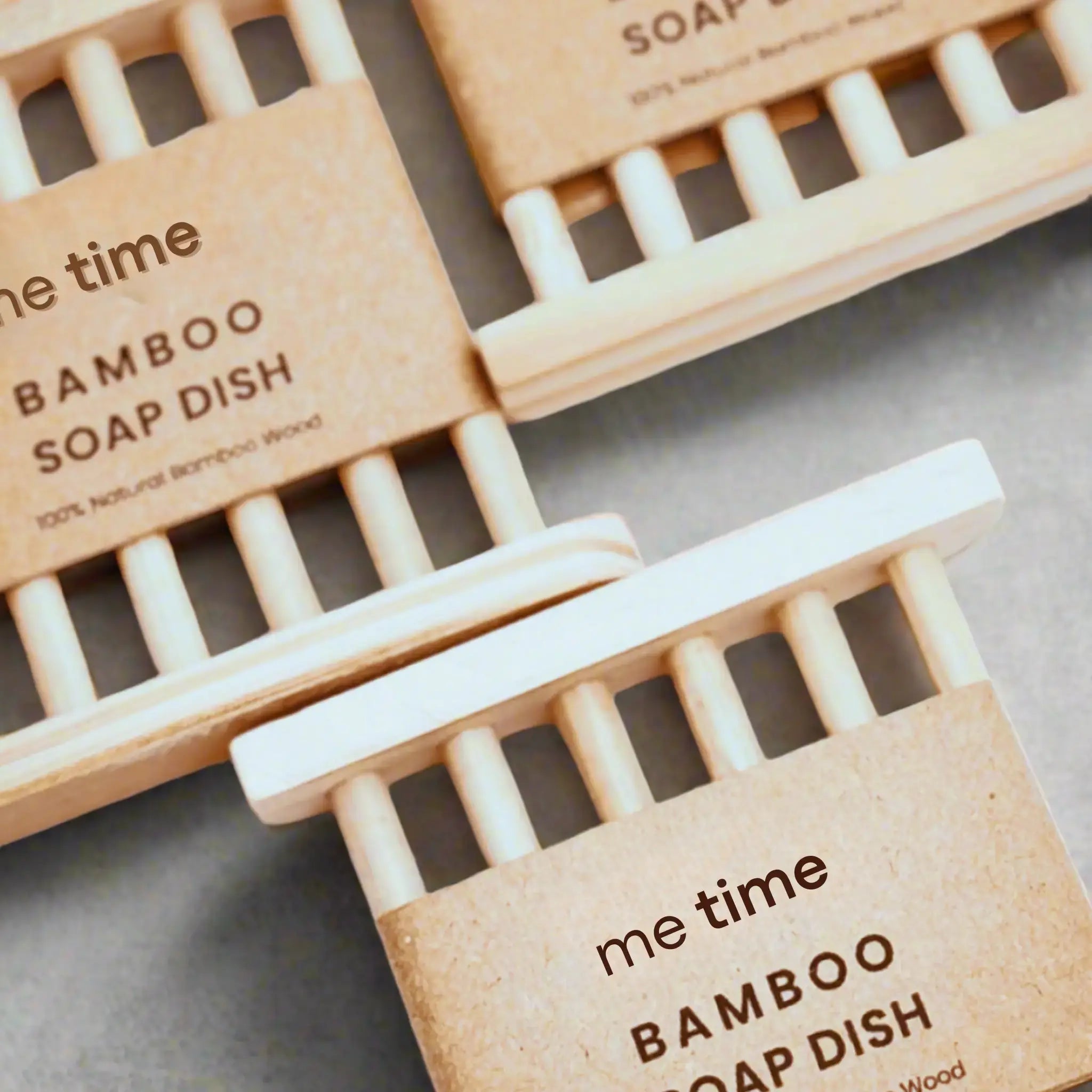 Three bamboo soap dishes with kraft paper labels arranged diagonally on a gray surface. The labels read 'me time BAMBOO SOAP DISH' and '100% Natural Bamboo Wood'.
