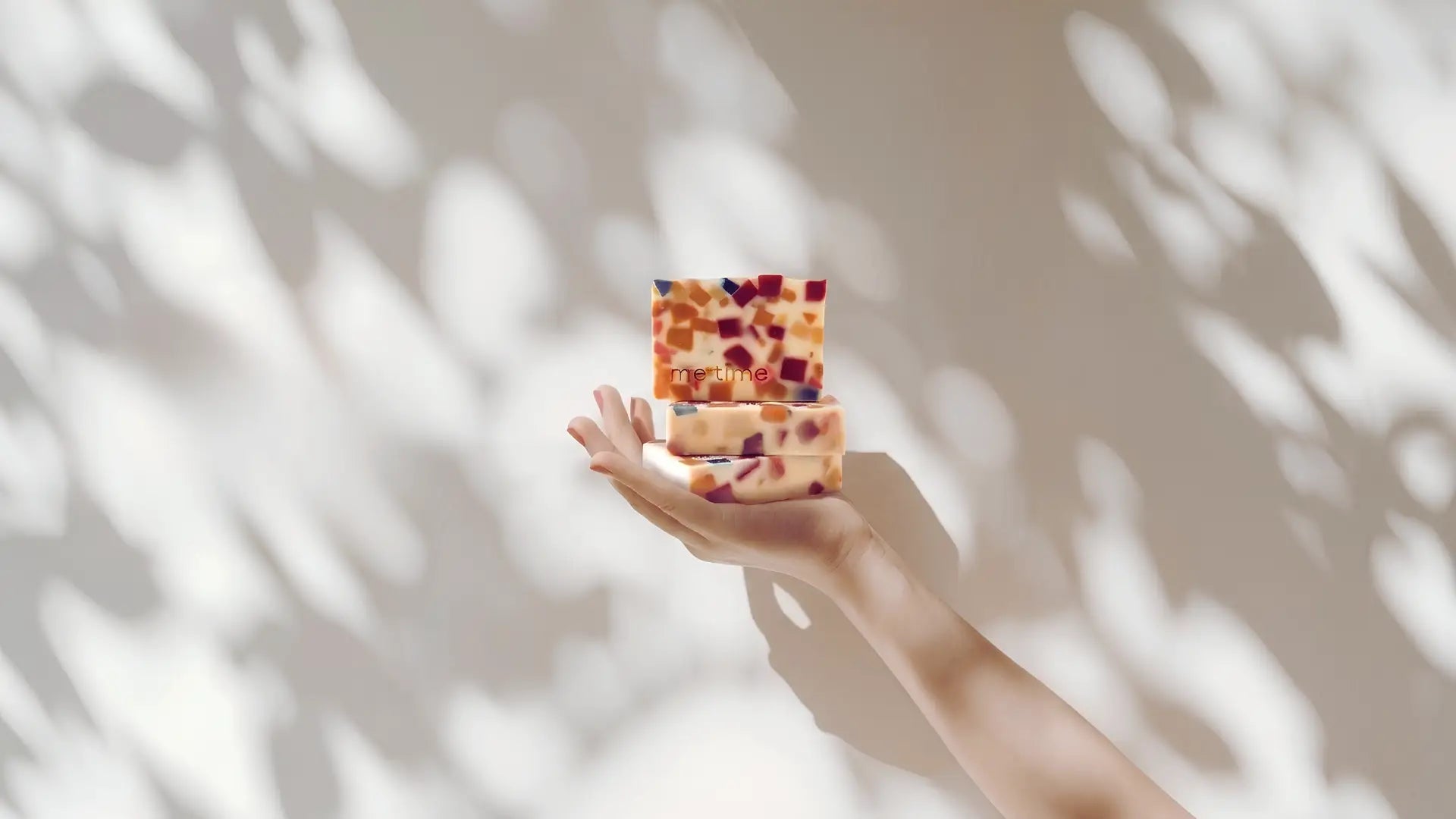 A hand holding two small rectangular bars of soap with a multicolor speckled pattern, set against a warm beige wall with soft leaf shadows.