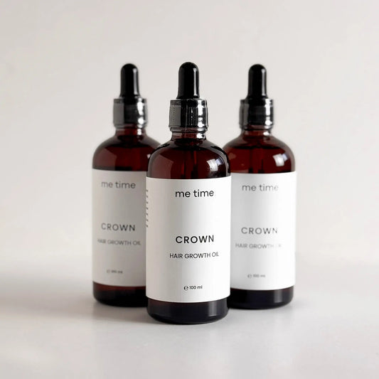 Elegant glass bottle of Crown Hair Growth Oil placed on a natural light grey surface, surrounded by fresh rosemary, black seeds, amla, and argan nuts, showcasing its premium natural ingredients for healthier, thicker hair.