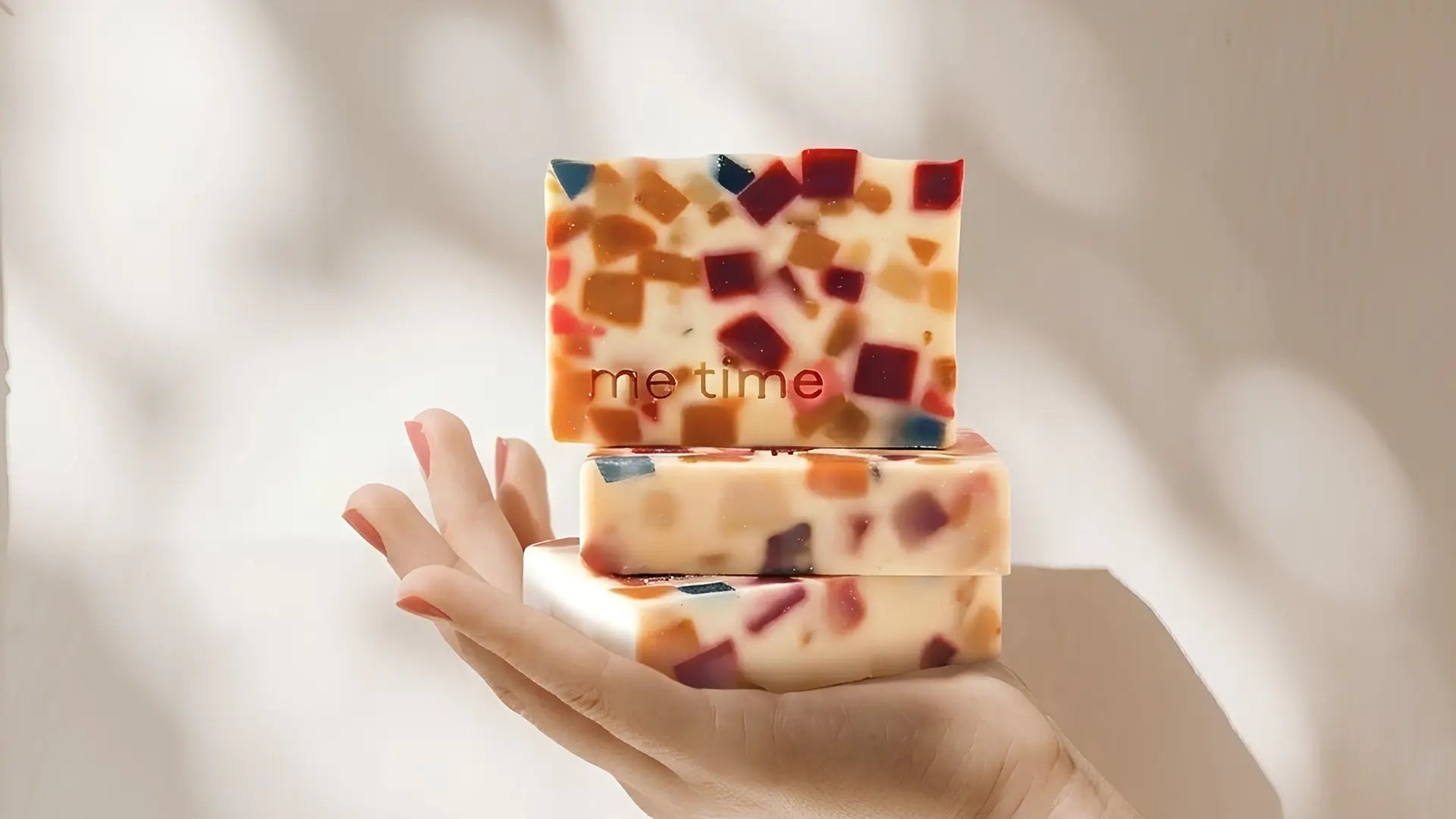 Three rectangular bars of handmade mosaic soap, stacked on a person's hand against a softly lit beige background with leaf shadows.