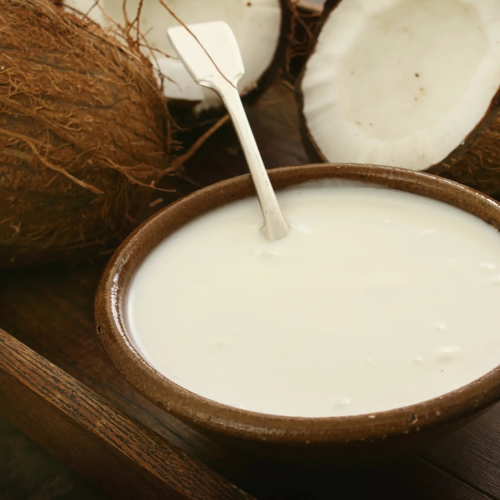 Coconut milk in a brown bowl with a spoon, surrounded by coconuts on a wooden surface.