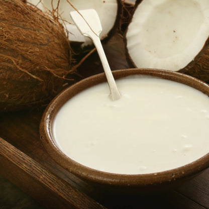 Coconut milk in a brown bowl with a spoon, surrounded by coconuts on a wooden surface.